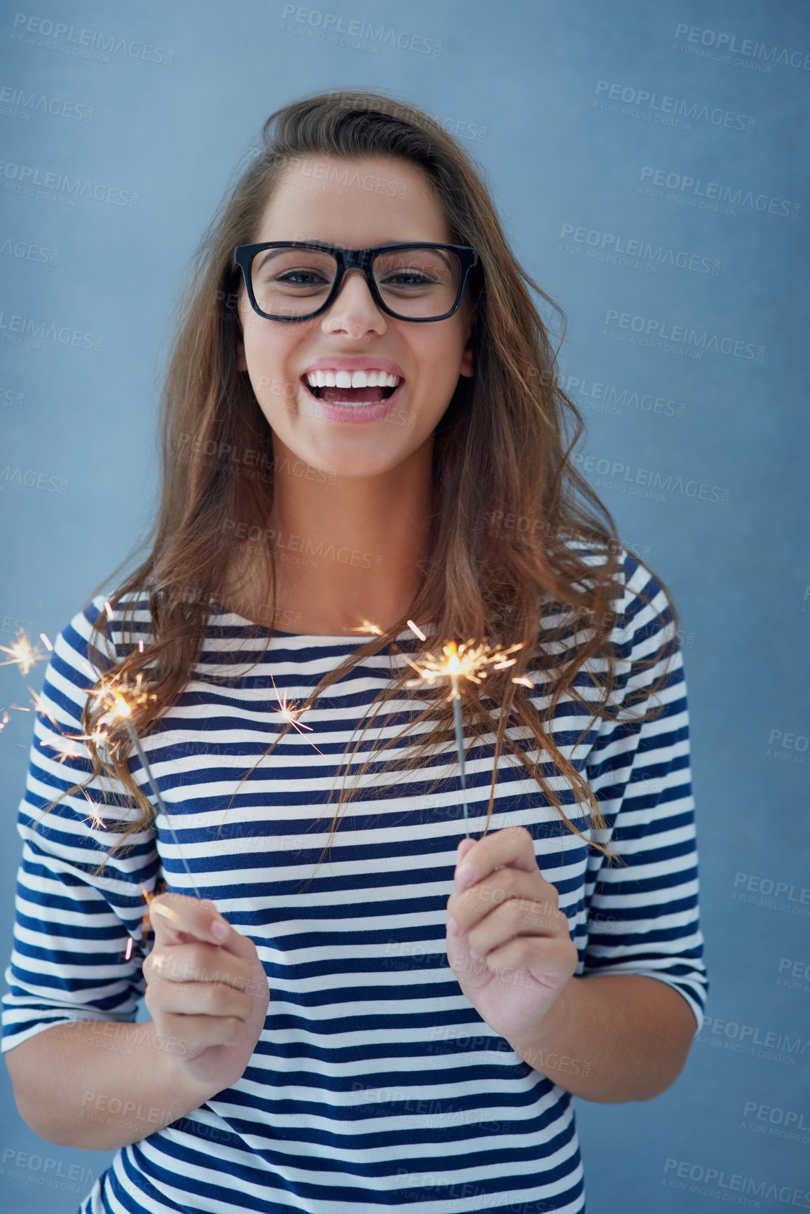 Buy stock photo Smile, portrait and woman in studio with sparklers for celebration isolated on blue background with light. Happy, glasses and fun event with birthday or new year with glow, vision and fireworks.