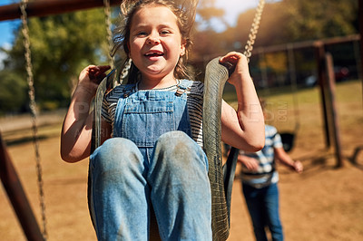 Buy stock photo Portrait of a little girl playing on a swing at the park with her brother in the background