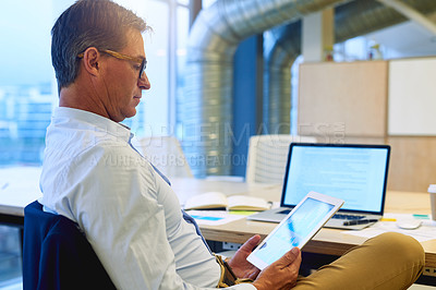 Buy stock photo Shot of a focussed businessman using his tablet while sitting at his desk in the office