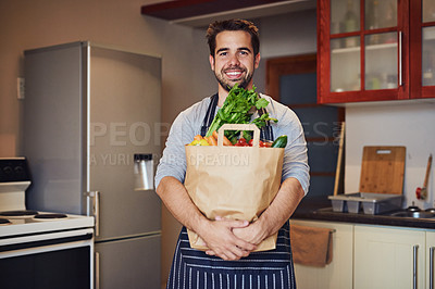 Buy stock photo Portrait of a happy young man posing with a bag of groceries in his kitchen at home
