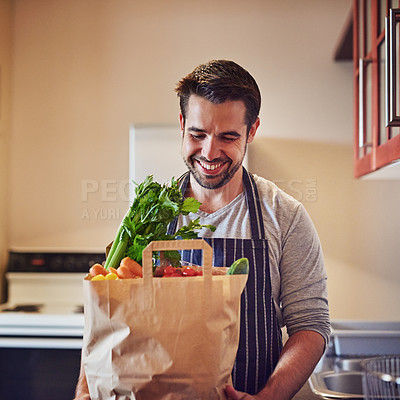 Buy stock photo Shot of a happy young man holding a bag of groceries in his kitchen at home