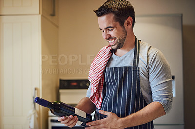 Buy stock photo Shot of a happy young man holding a bottle of wine while standing in his kitchen at home