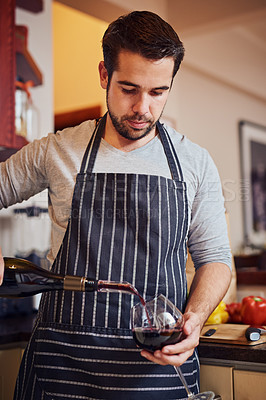 Buy stock photo Shot of a happy young man pouring himself a glass of wine while cooking in his kitchen at home