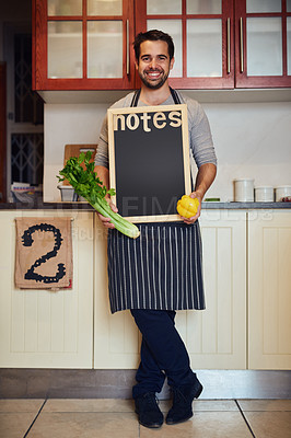 Buy stock photo Portrait of a happy young man posing with fresh ingredients and a blank chalkboard in his kitchen