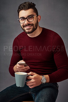 Buy stock photo Studio portrait of a handsome young man texting on his cellphone while drinking coffee against a dark background