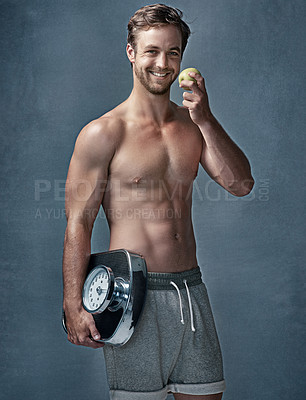 Buy stock photo Portrait of a healthy young man eating an apple while posing with a scale in studio