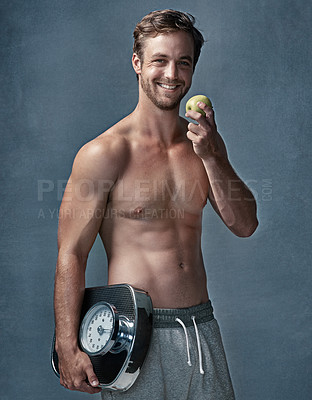Buy stock photo Portrait of a healthy young man eating an apple while posing with a scale in studio