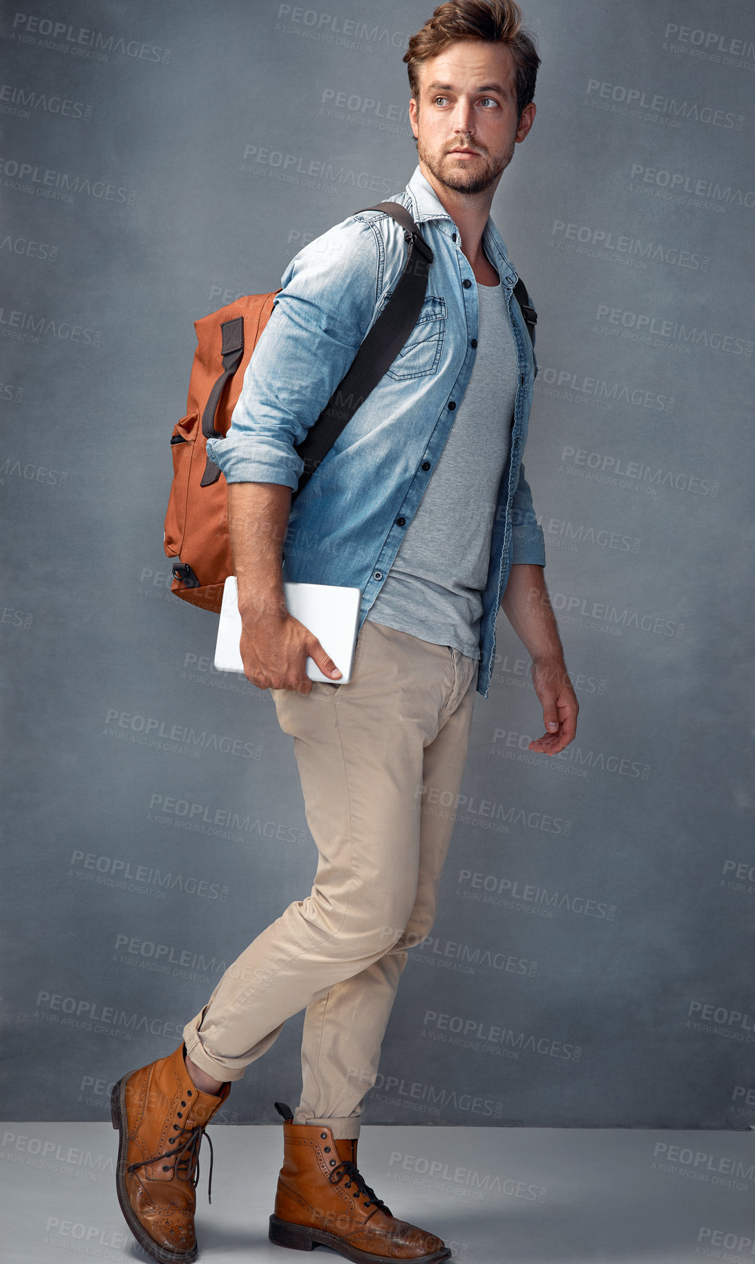 Buy stock photo Shot of a handsome young man  with a backpack holding a tablet while standing in the studio
