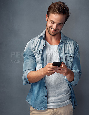 Buy stock photo Shot of a happy young man using his smartphone while standing against a gray background in the studio