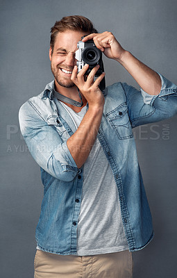 Buy stock photo Portrait of a happy young man holding up a camera while posing against a gray background in the studio