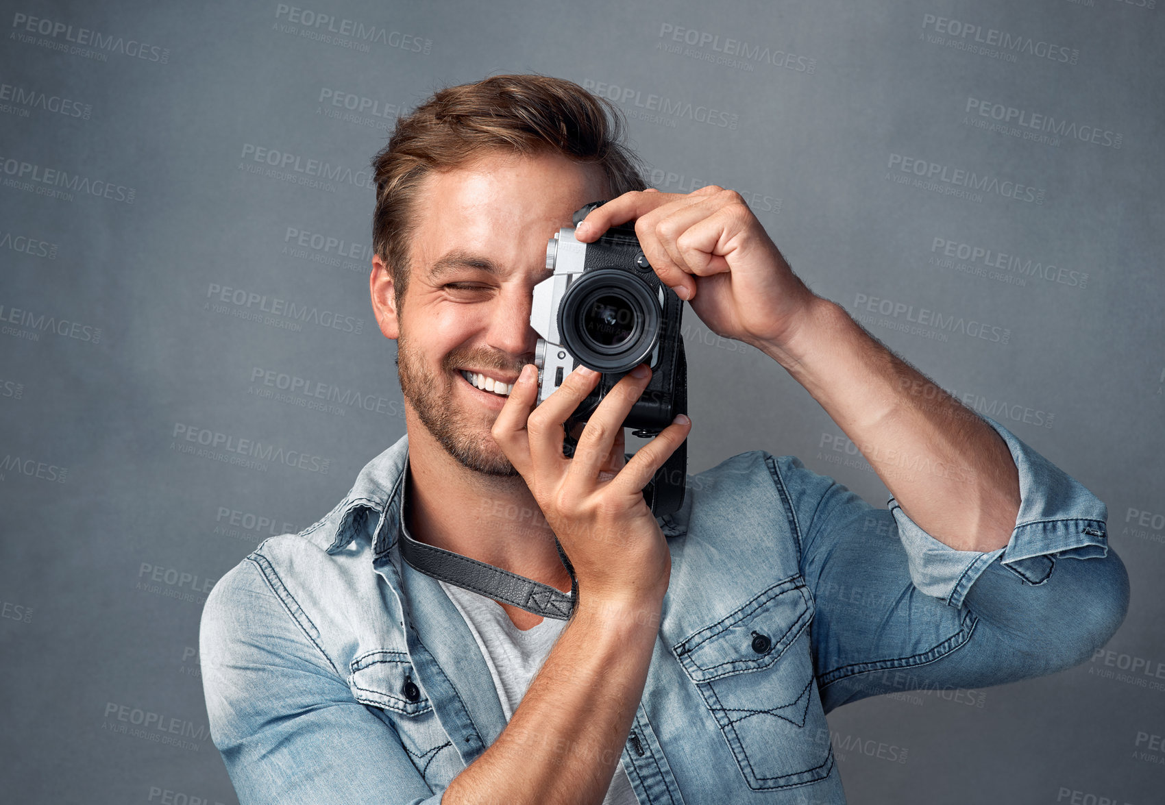 Buy stock photo Portrait of a happy young man holding up a camera while posing against a gray background in the studio