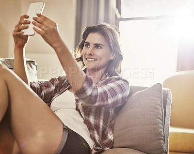 Buy stock photo Shot of a young woman relaxing on the sofa at home and using a mobile phone to take selfies
