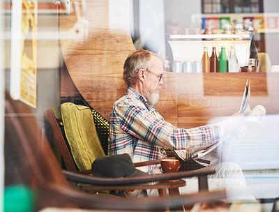 Buy stock photo Shot of a senior man through a window reading the newspaper at a coffee shop