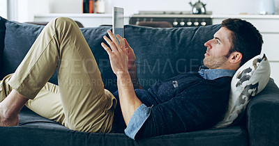 Buy stock photo Cropped shot of a handsome young man using a digital tablet while lying on the sofa at home
