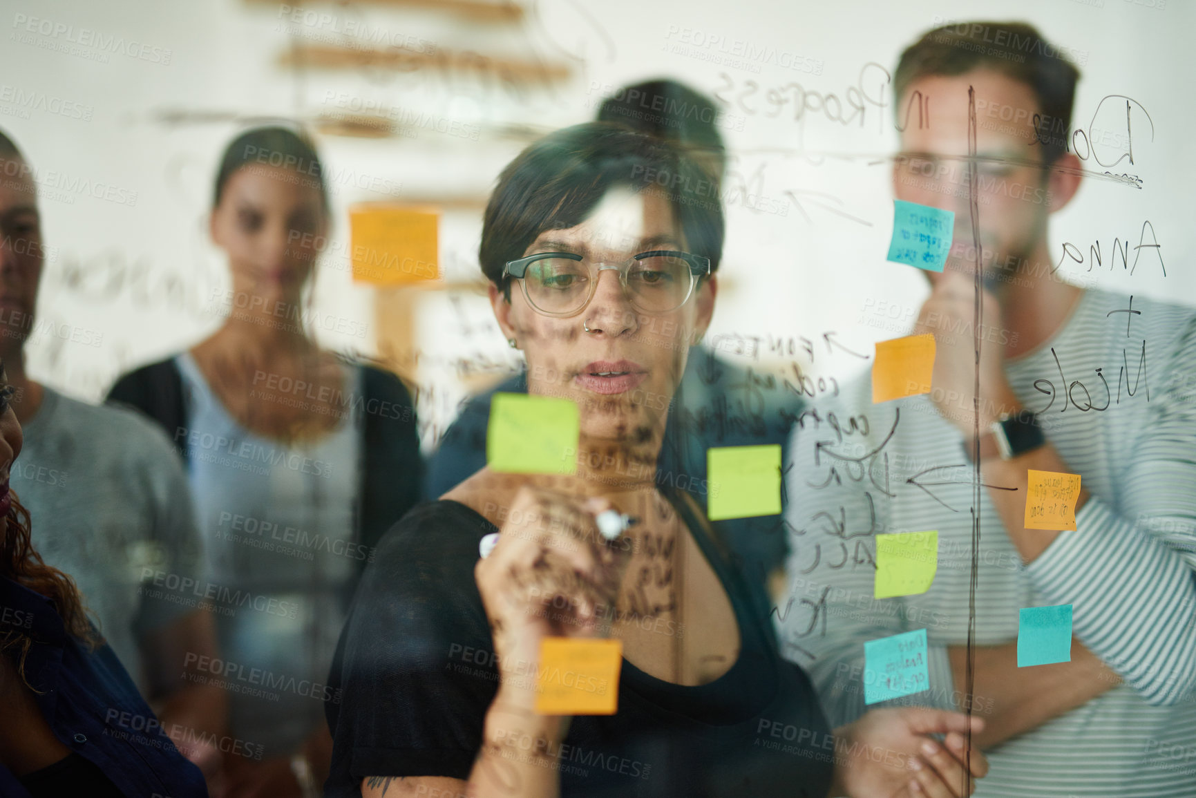 Buy stock photo Professional, smart and creative businesswoman brainstorming a strategy idea and writing on sticky notes on a transparent board with colleagues. Diverse group planning and working together in a team