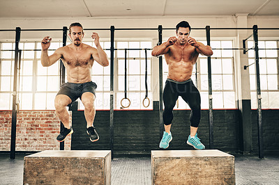 Buy stock photo Shot of two fit and determined young men jumping and doing squats to workout in a gym
