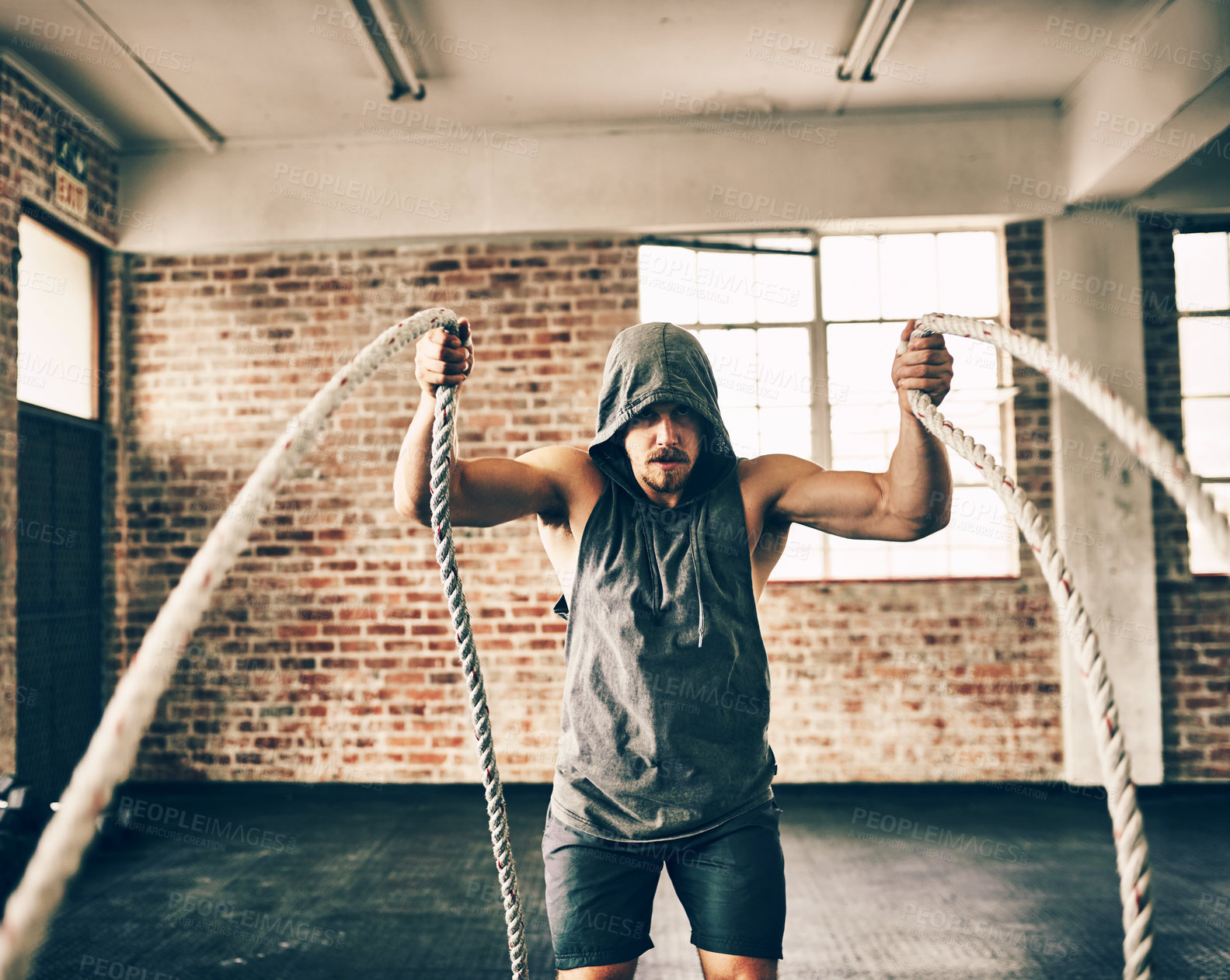 Buy stock photo Shot of a hooded and determined young man making use of ropes to workout in the gym