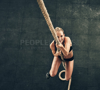 Buy stock photo High angle shot of a fit and determined young woman climbing a rope for strength exercise in a gym