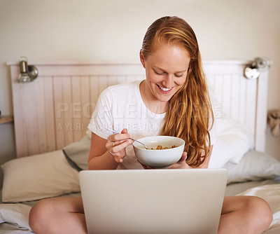 Buy stock photo Shot of a young woman eating breakfast and using a laptop at home