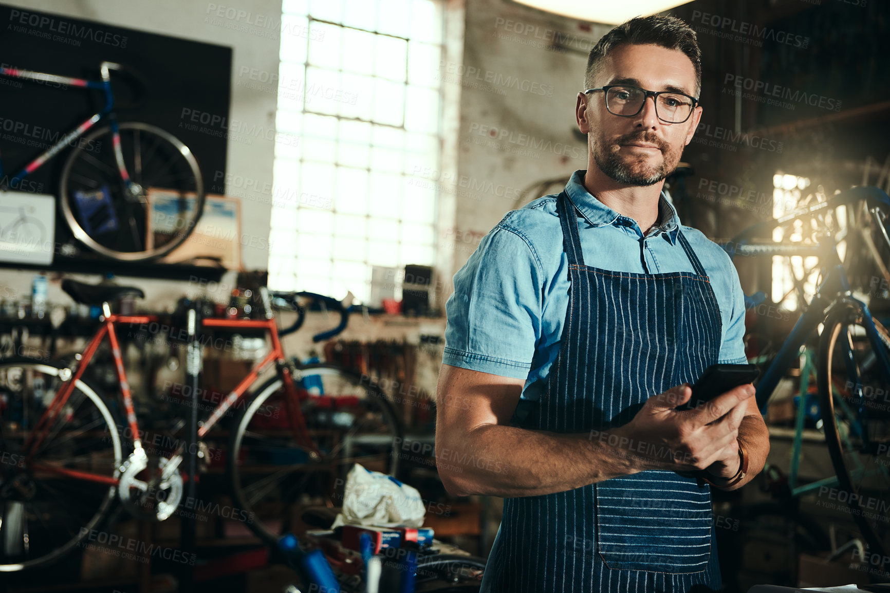 Buy stock photo Bicycle, garage and phone for man, portrait and small business of mechanic, apron and equipment. Proud, owner and person with glasses, maintenance and tools for bike, technician or mobile for service