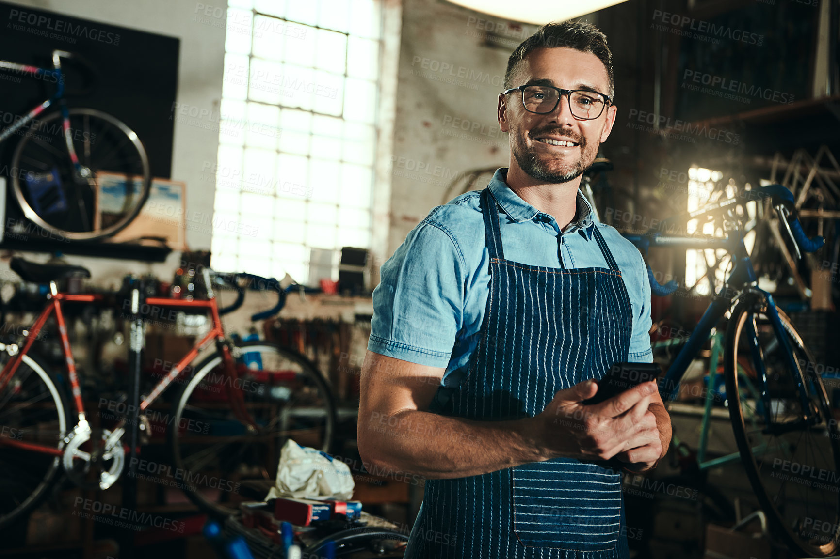 Buy stock photo Bicycle, small business and phone of man, smile and garage of mechanic, apron and equipment. Portrait, owner and person with service, maintenance and tools for bike, technician or working with mobile