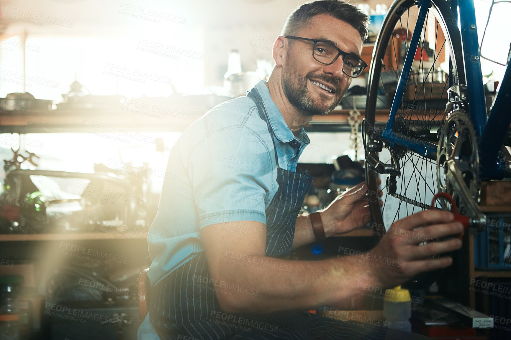 Buy stock photo Man, garage and confident as bicycle mechanic with lens flare in startup or small business. Workshop, portrait and smile or happy as handyman with fixing bike at repair or maintenance shop and proud