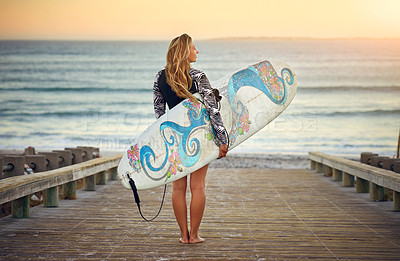 Buy stock photo Rearview shot of an attractive young female surfer standing with her surfboard looking over the ocean