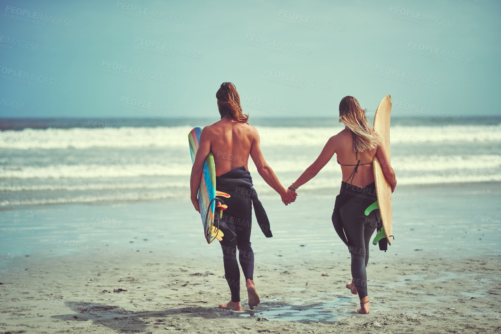 Buy stock photo Rearview shot of a young couple walking on the beach with their surfboards