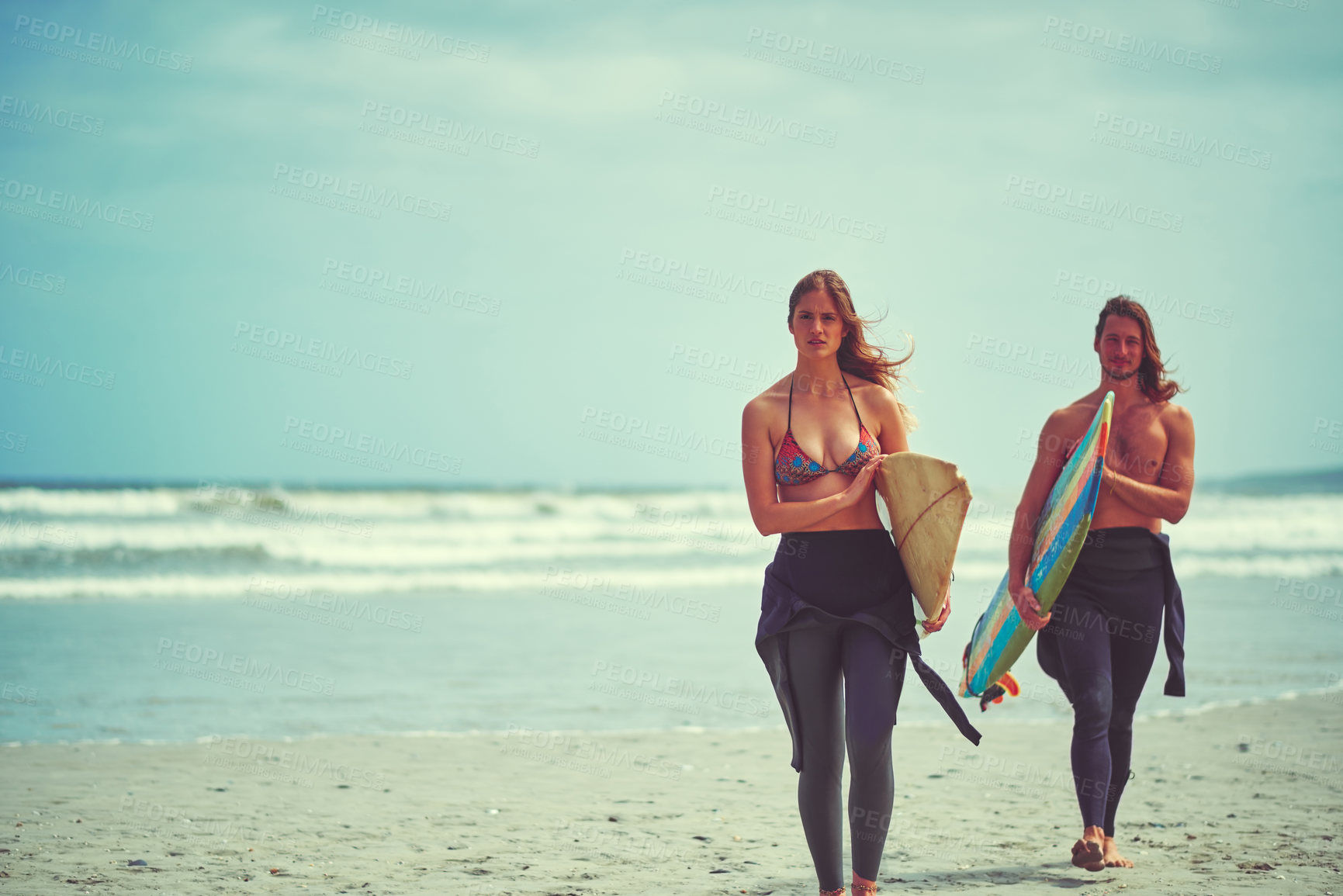Buy stock photo Shot of a young couple walking on the beach with their surfboards
