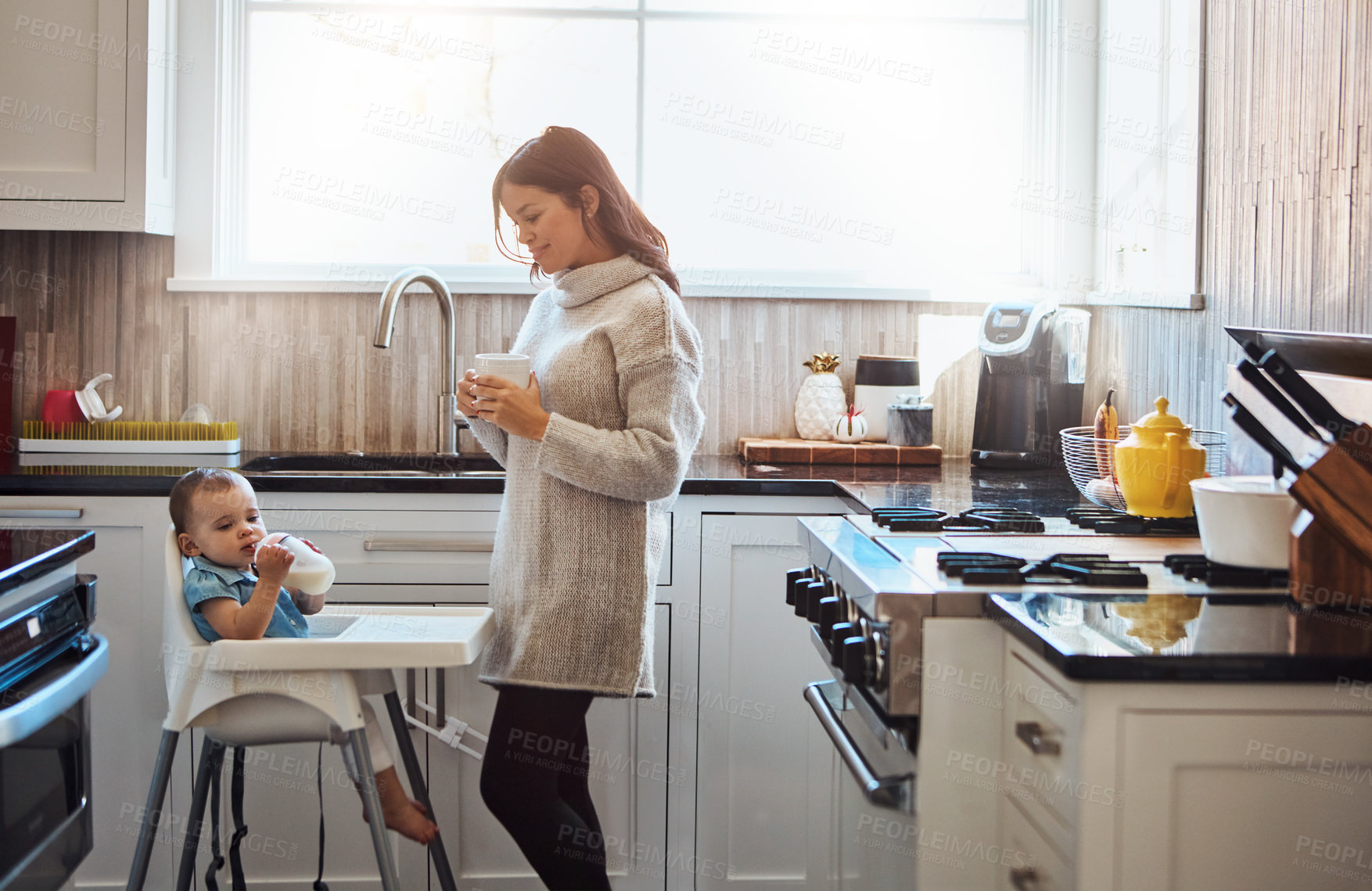 Buy stock photo Shot of an adorable baby girl drinking bottled milk in her high chair while her mother has coffee