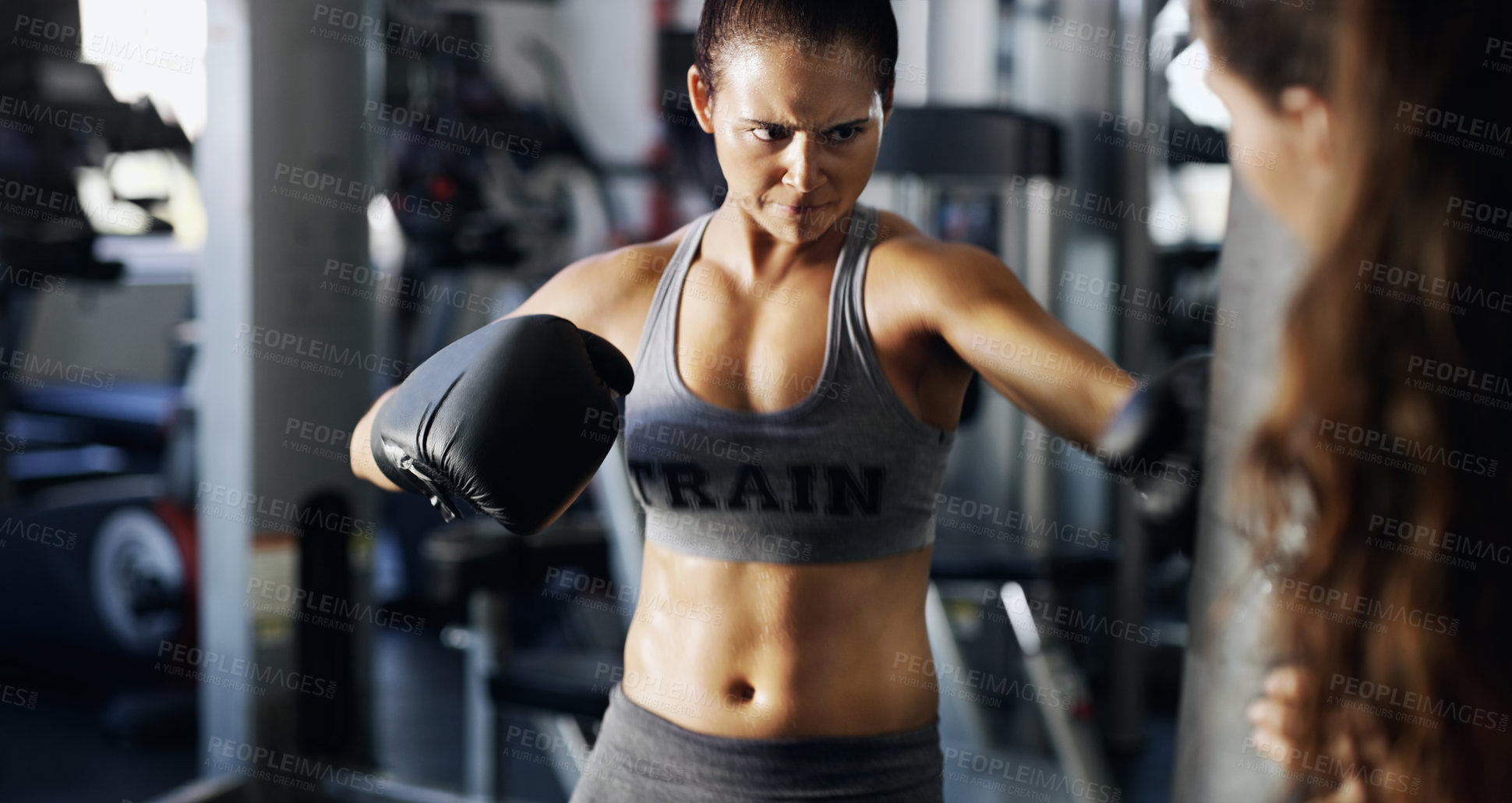 Buy stock photo Cropped shot of a young female boxer working out on a punching bag with her trainer in the gym