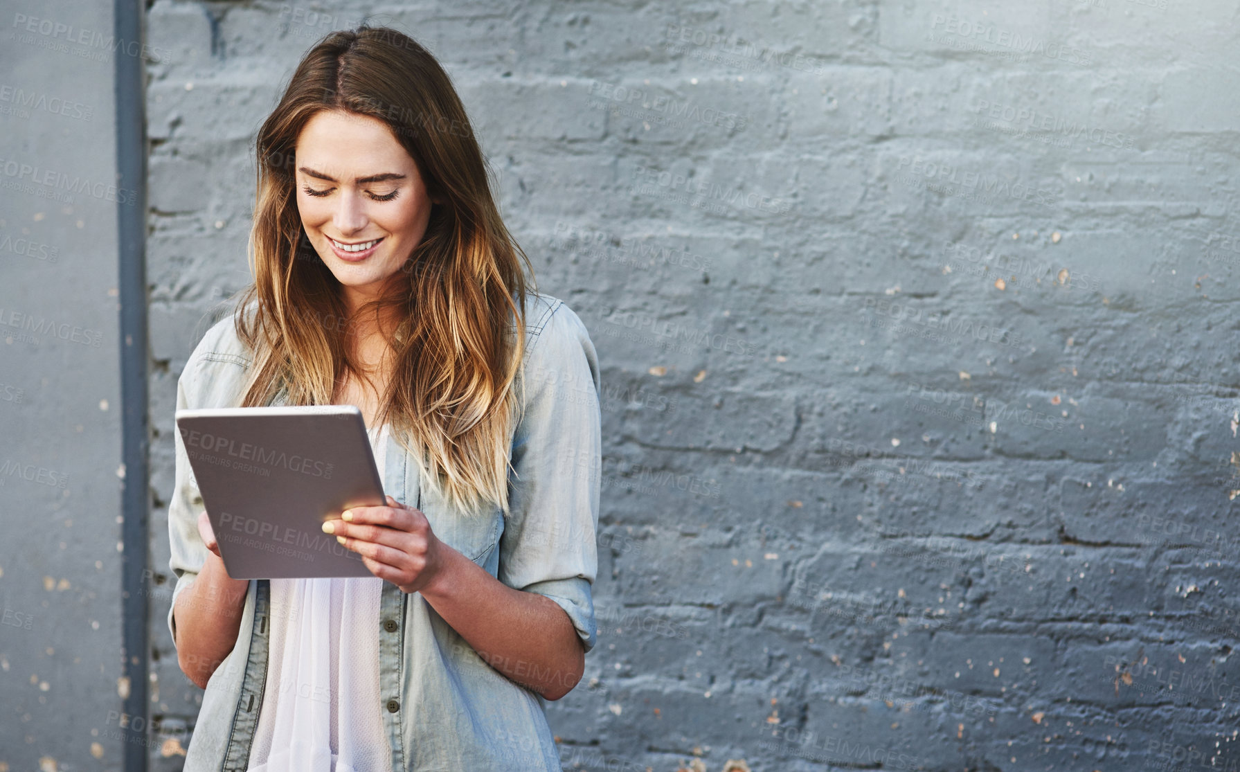 Buy stock photo Shot of a young woman standing outdoors and using a digital tablet against a gray wall