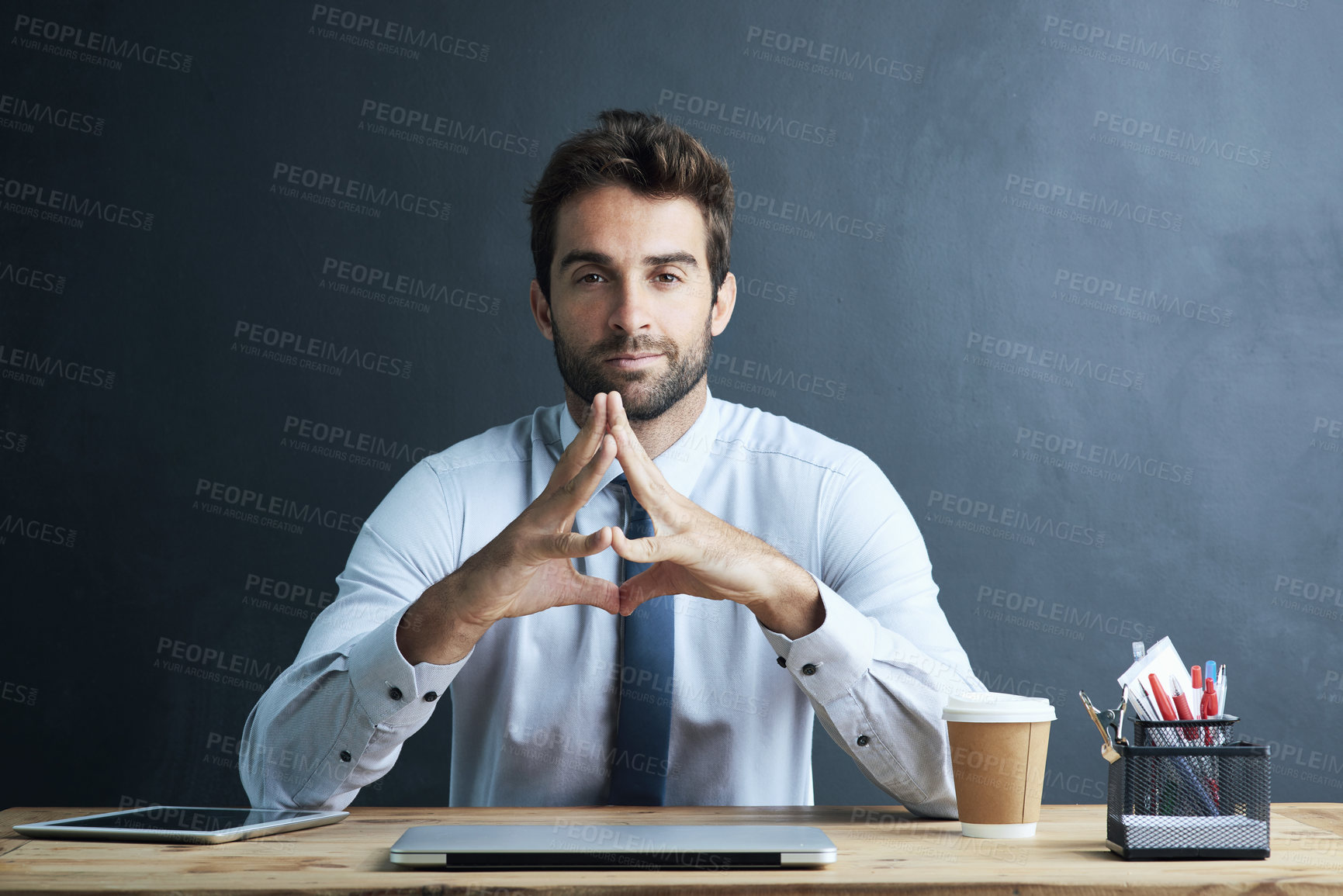 Buy stock photo Portrait of a young corporate businessman sitting at a desk against a dark background