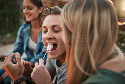 Buy stock photo Shot of a young cheerful man being fed a marshmallow by his girlfriend while being seated outside
