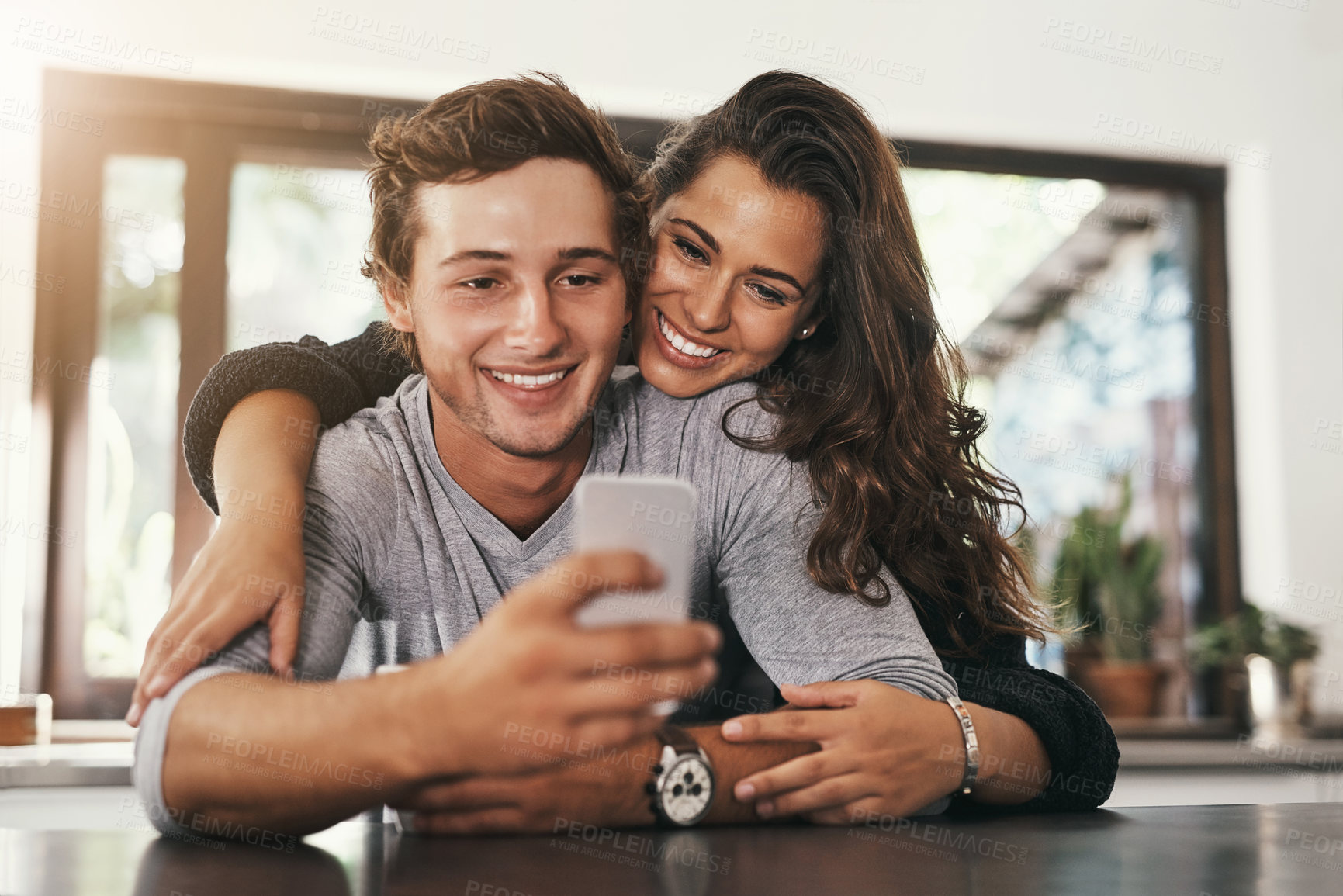 Buy stock photo Couple, relax and hug with phone in kitchen for network, communication or post on social media. Man, woman and smile with tech in morning for text message, notification or reading online news at home
