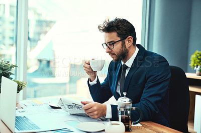 Buy stock photo Shot of a young businessman drinking a cup of tea while reading a newspaper in an office