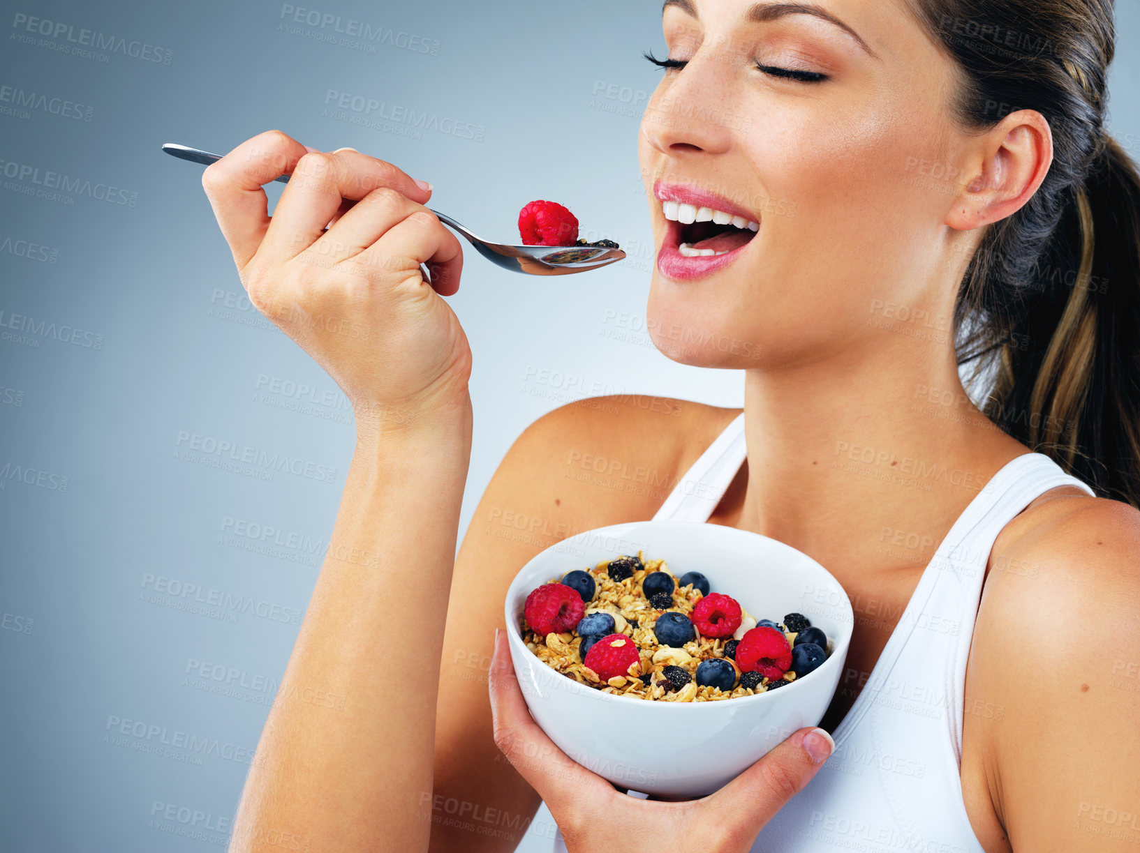 Buy stock photo Happy woman, eating and bowl with fruit for diet or nutrition on a blue studio background. Young female person or model with smile or spoon for natural organic muesli, healthy wheat or vitamin fiber