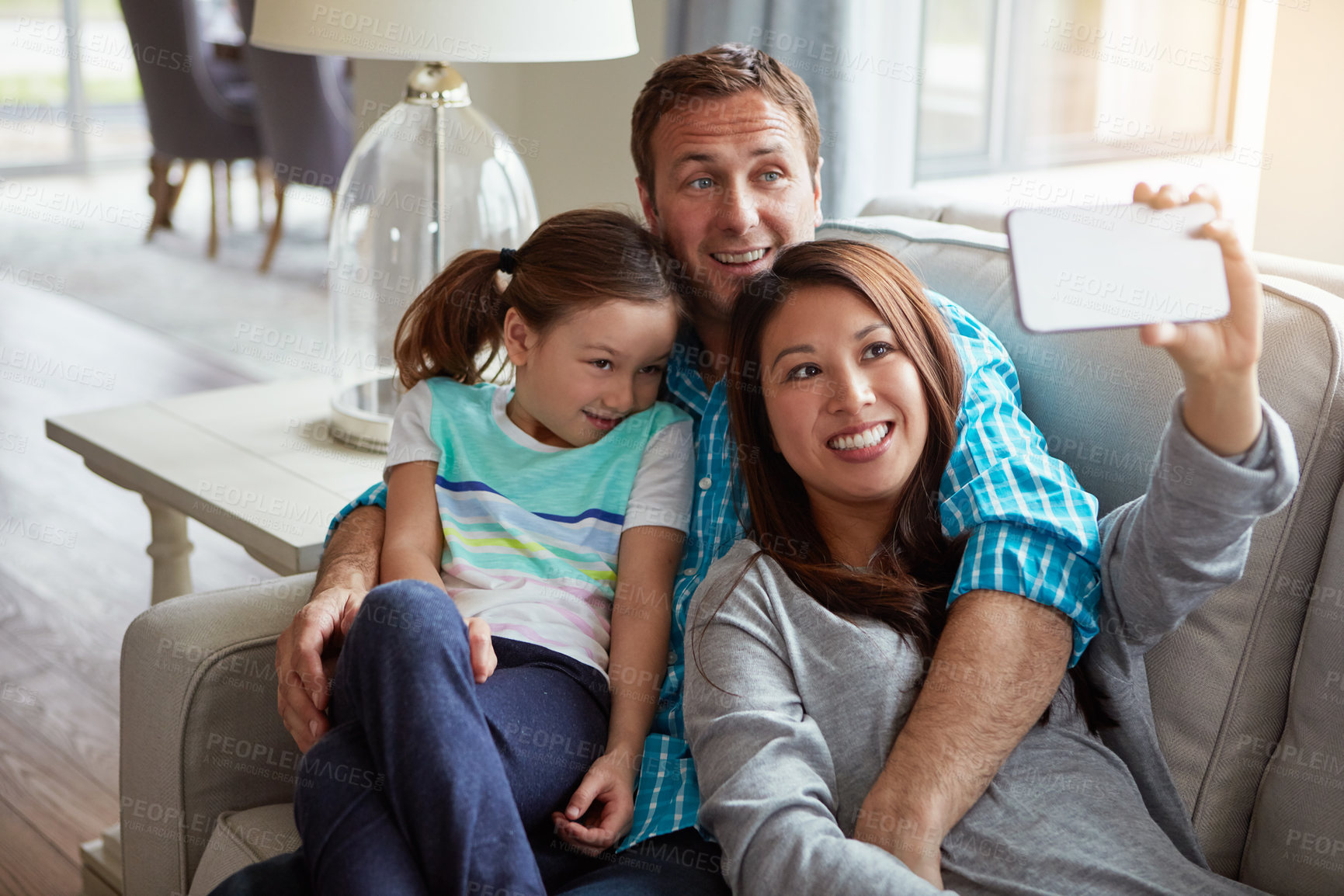 Buy stock photo Interracial parents, girl and selfie on couch for smile, hug and relax with care, mobile app or memory in home. Father, mother and daughter with embrace, social media or love on sofa in family house