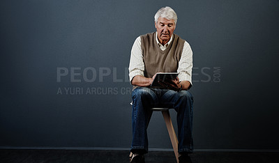 Buy stock photo Studio shot of a handsome mature man using a tablet while sitting on a wooden stool against a dark background