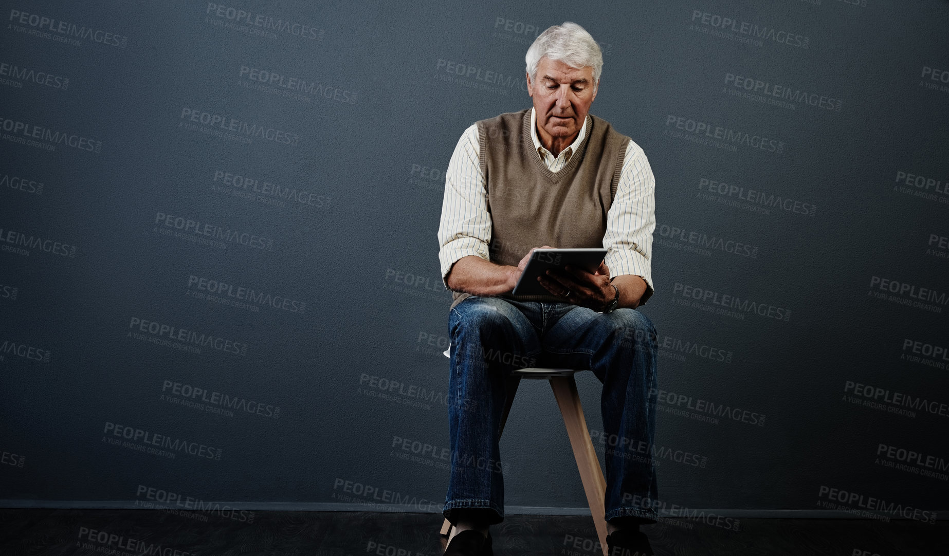 Buy stock photo Studio shot of a handsome mature man using a tablet while sitting on a wooden stool against a dark background