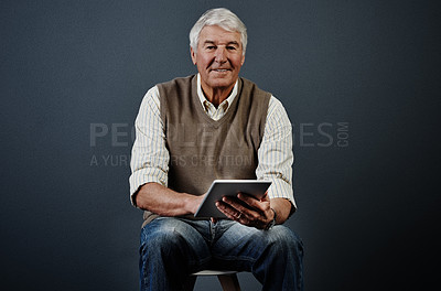 Buy stock photo Studio portrait of a handsome mature man using a tablet while sitting on a wooden stool against a dark background