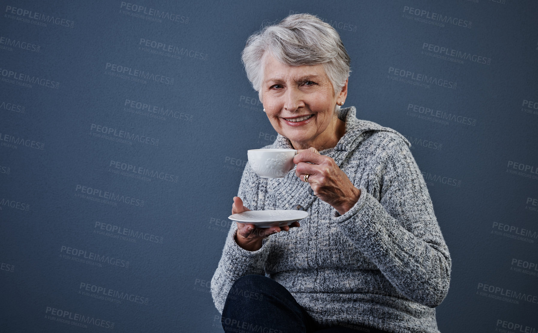 Buy stock photo Studio shot of a cheerful elderly woman sitting down and enjoying a cup of tea while looking at the camera