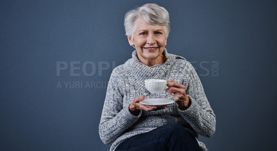 Buy stock photo Studio shot of a cheerful elderly woman sitting down and enjoying a cup of tea while looking at the camera