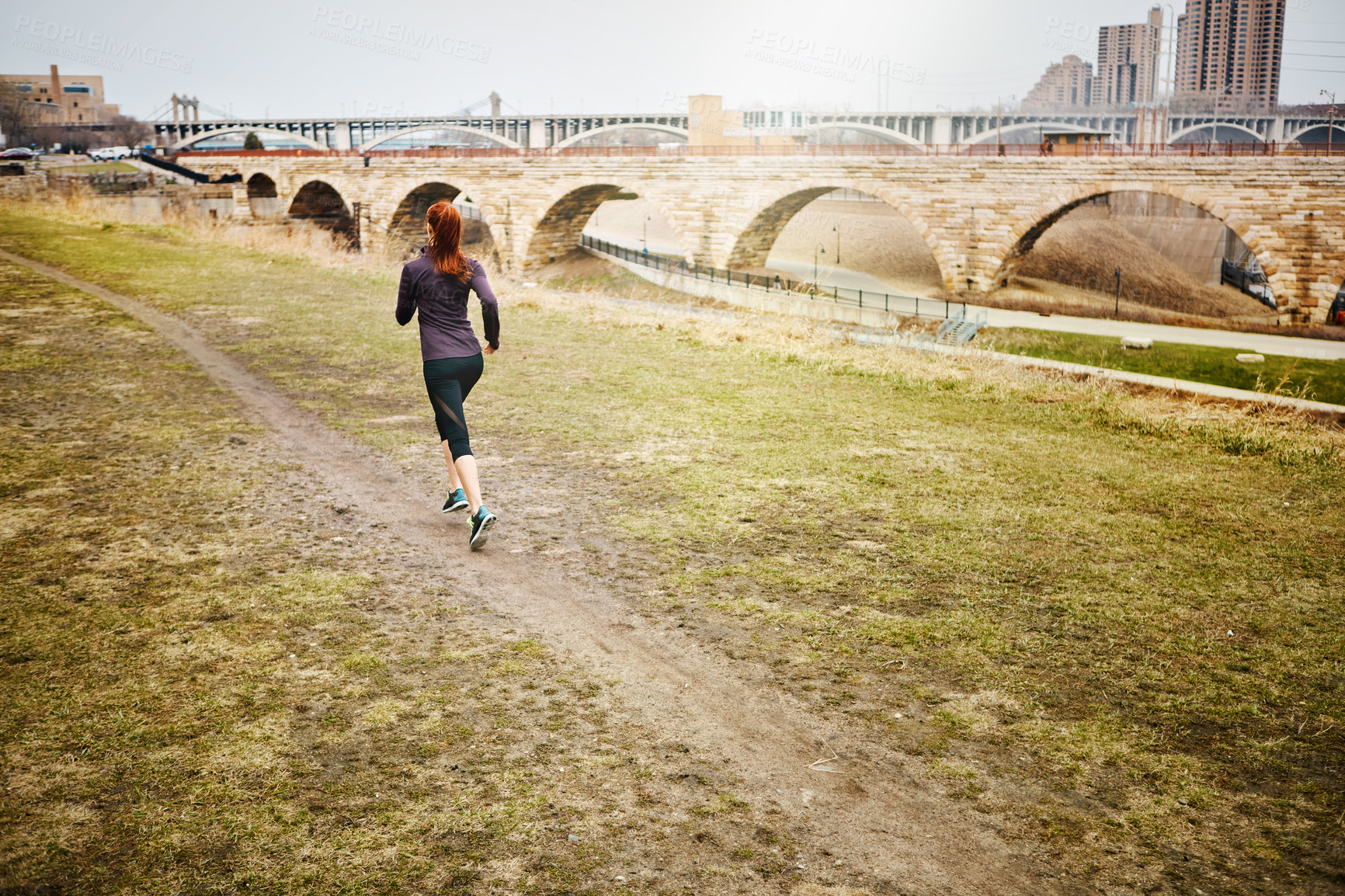 Buy stock photo Rear view shot of a sporty young woman running alongside the city