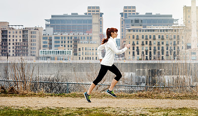 Buy stock photo Shot of a sporty young woman running alongside the city