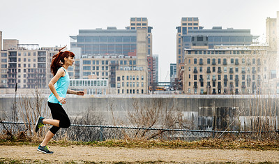 Buy stock photo Shot of a sporty young woman running alongside the city