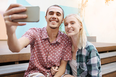 Buy stock photo Shot of a happy young couple using a mobile phone to take selfies outside