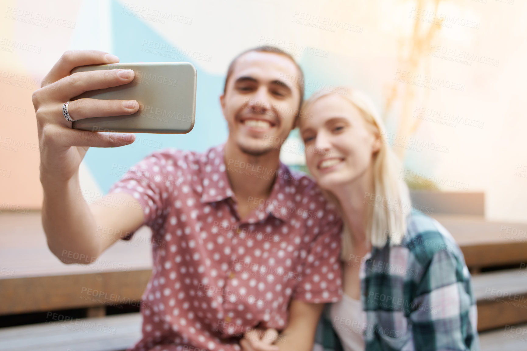 Buy stock photo Shot of a happy young couple using a mobile phone to take selfies outside