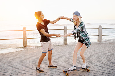 Buy stock photo Full length shot of a handsome young man teaching his girlfriend how to skateboard on the promenade