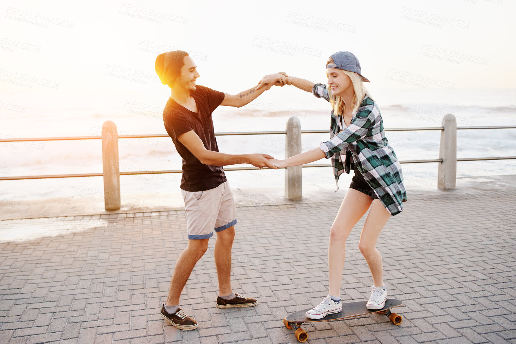 Buy stock photo Full length shot of a handsome young man teaching his girlfriend how to skateboard on the promenade